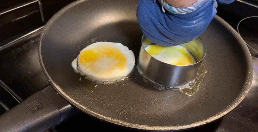 Fried egg cooking in a ring mold on a skillet, with a hand holding a metal ring above it.