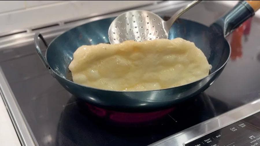 Golden-brown beavertail frying in a dark wok with a slotted spoon pressing it down in hot oil.
