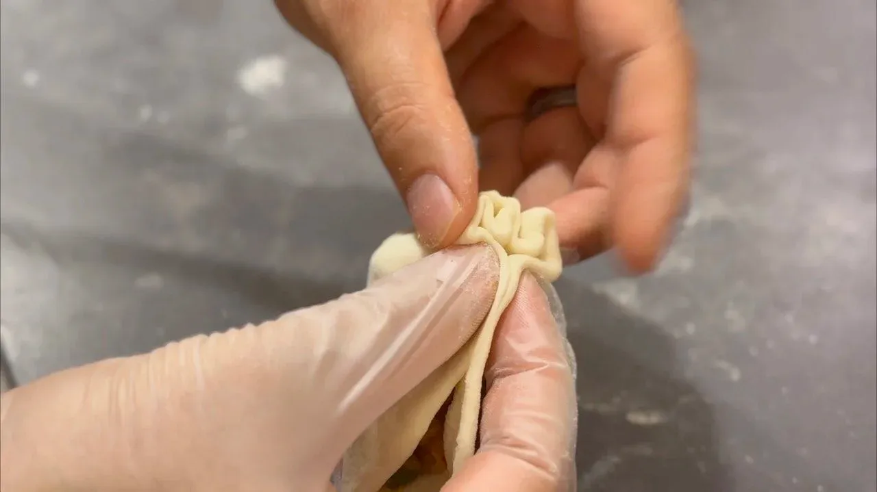 Hands pinching and twisting the top of a momo wrapper to seal it closed.