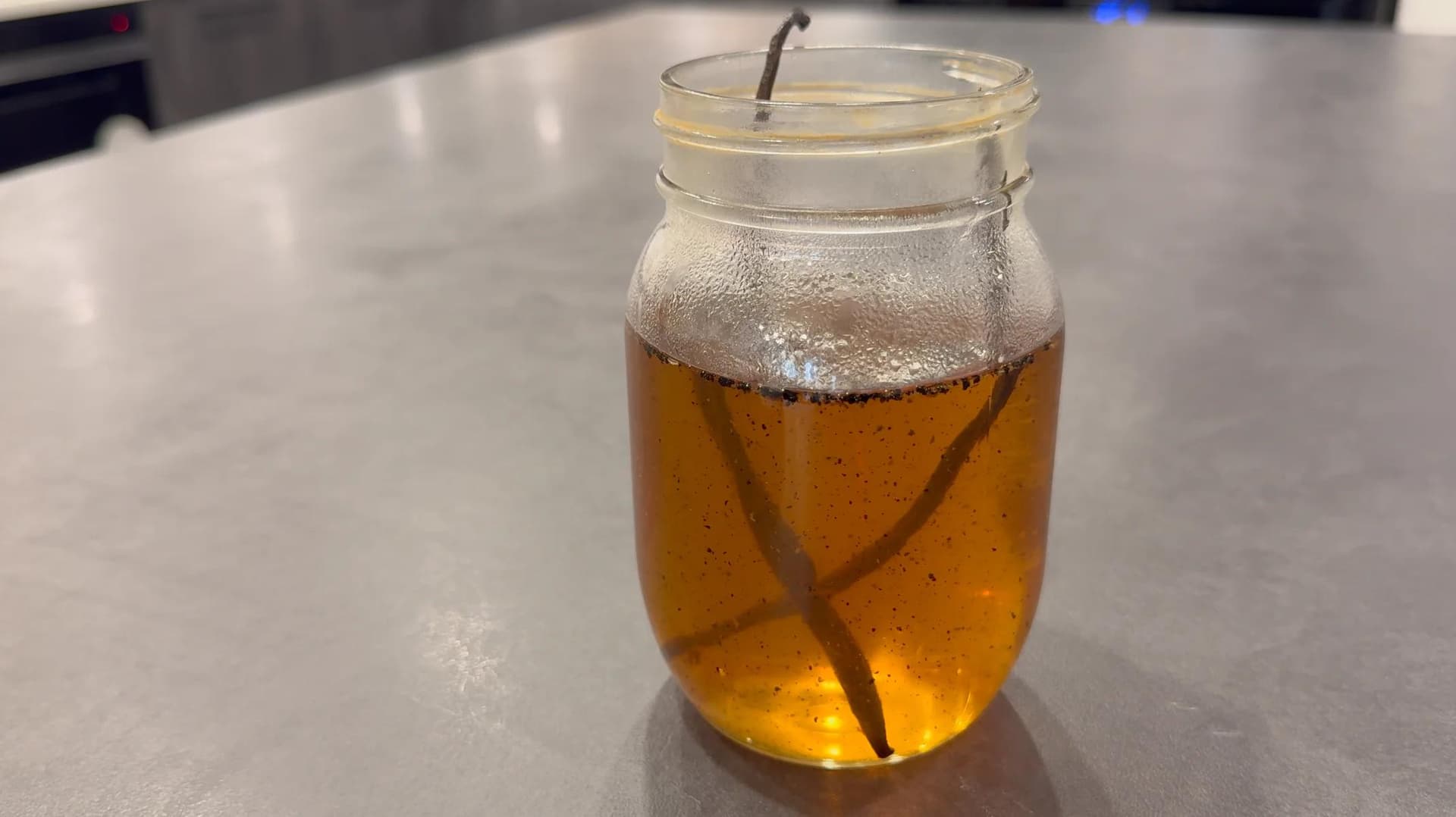 Vanilla syrup stored in a glass jar with vanilla bean pod, golden-brown liquid with condensation on the outside.
