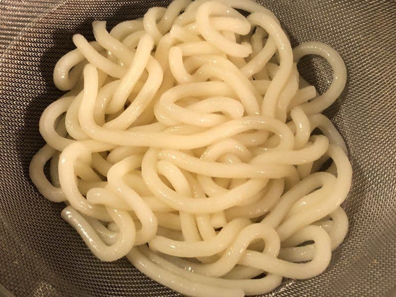 Cooked udon noodles drained in a colander, showing thick, curly strands of noodles.