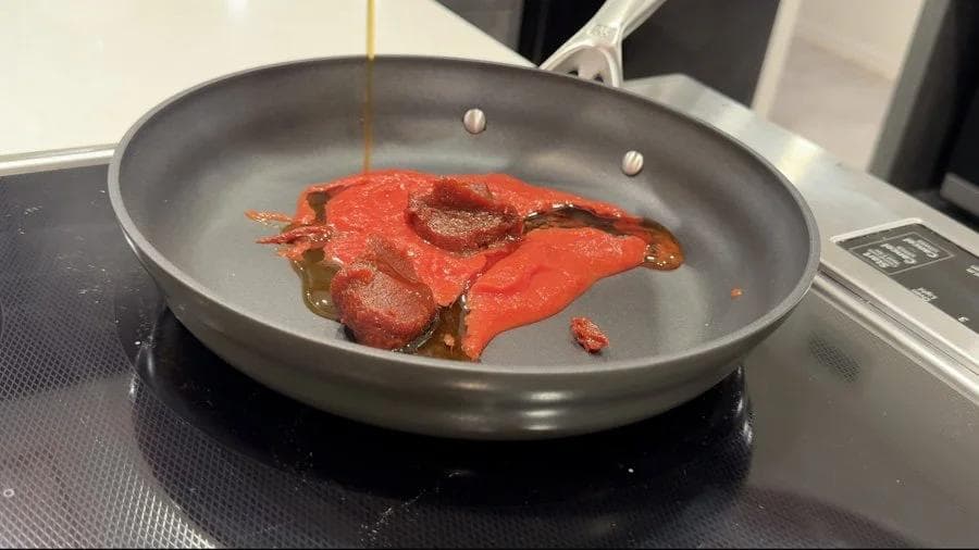 Red sauce simmering in a gray nonstick pan on a stovetop, with oil being drizzled in from above.