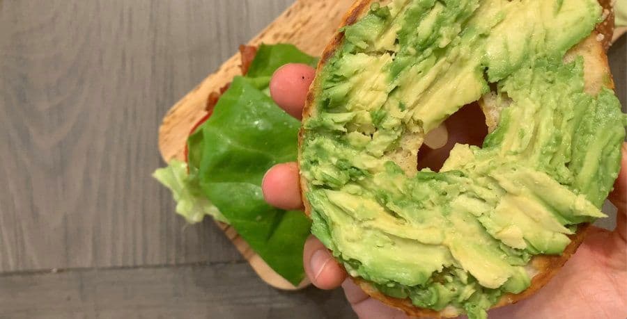 Hand holding a toasted bagel half topped with mashed avocado, lettuce, and other sandwich ingredients.