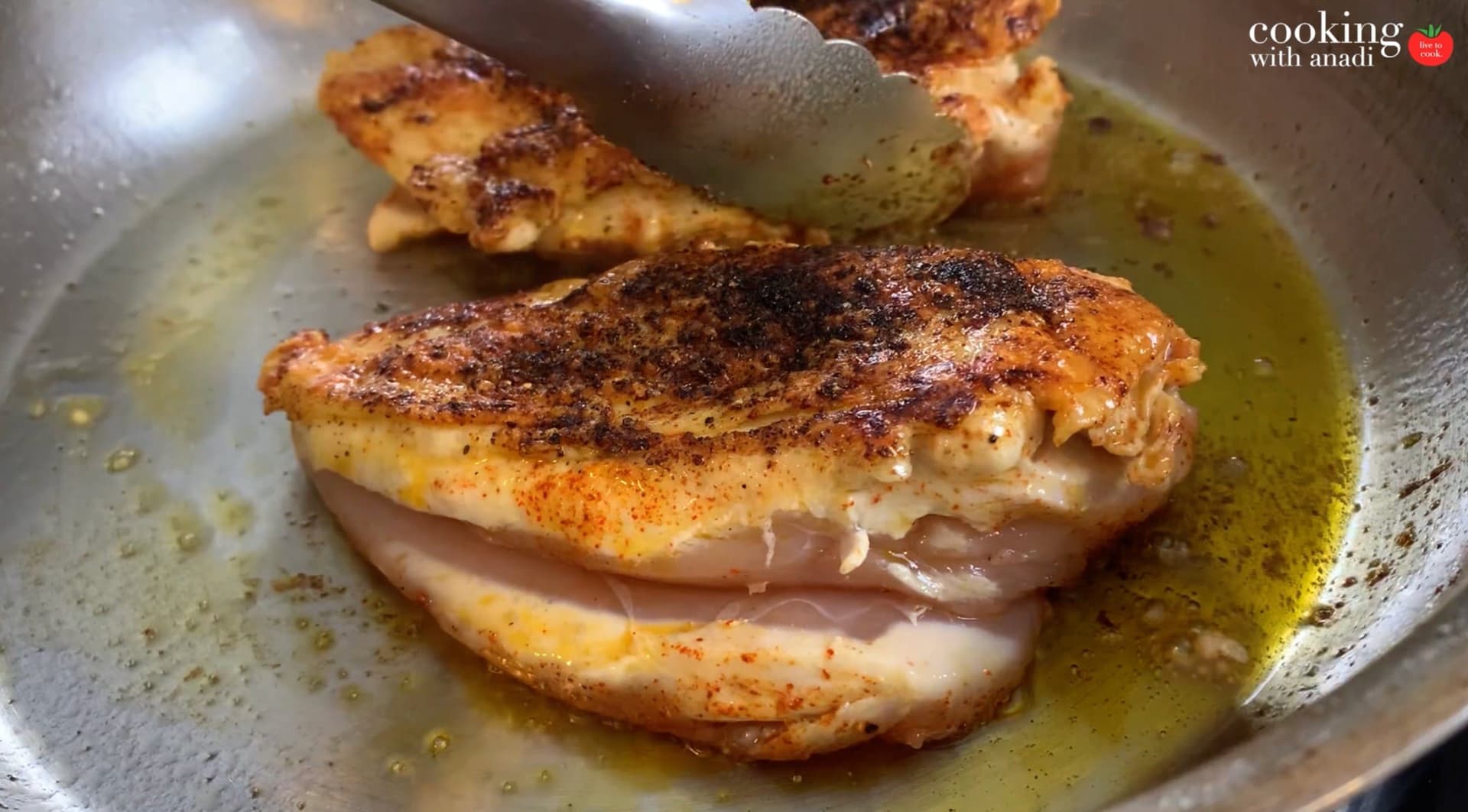 Seasoned chicken breasts searing in olive oil in a stainless steel pan, golden brown with spices.