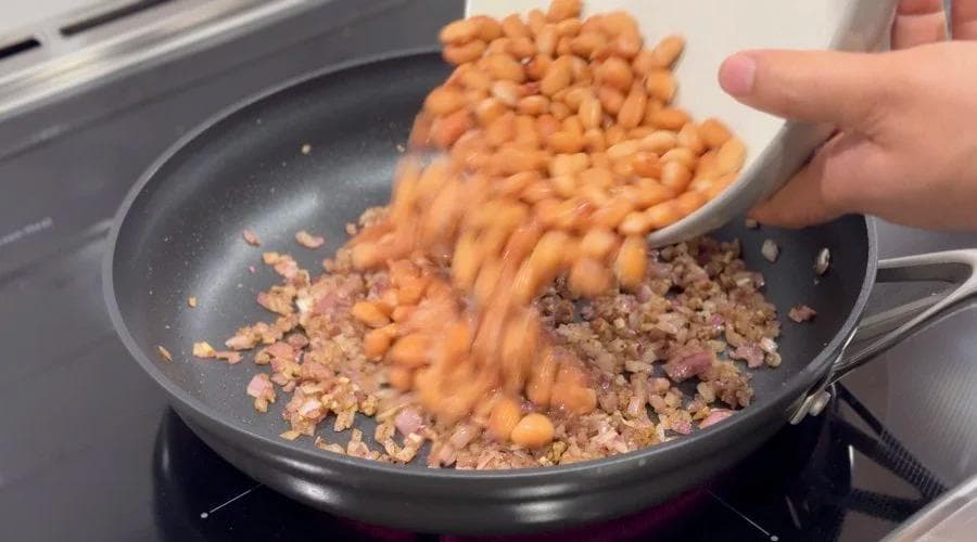 Canned beans being poured into a skillet with cooked ground meat and onions during preparation of refried beans.