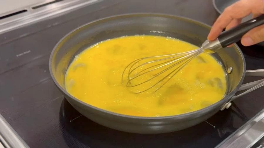 Whisking scrambled eggs in a skillet on the stovetop, eggs still slightly runny and pale yellow.