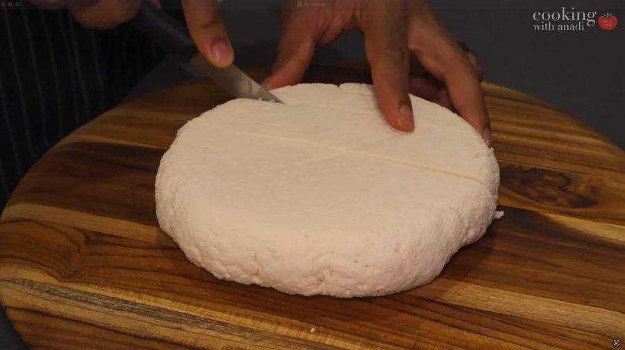 Hands pressing down on a block of fresh white paneer cheese on a wooden cutting board.