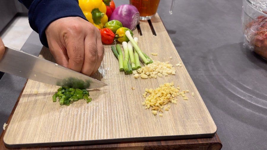 Chef's hands chopping garlic and green onions on cutting board with prepped bell peppers and green onions nearby.