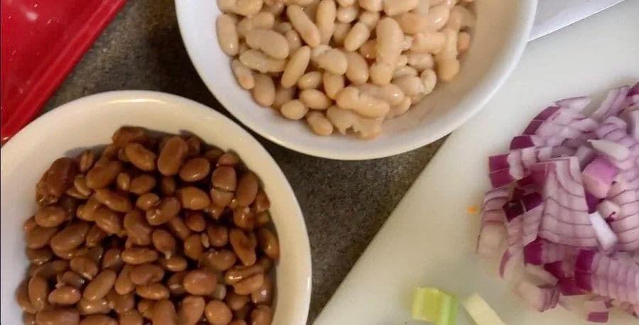 Ingredients for minestrone soup: cooked beans, white beans, and diced red onions in white bowls on a gray surface.
