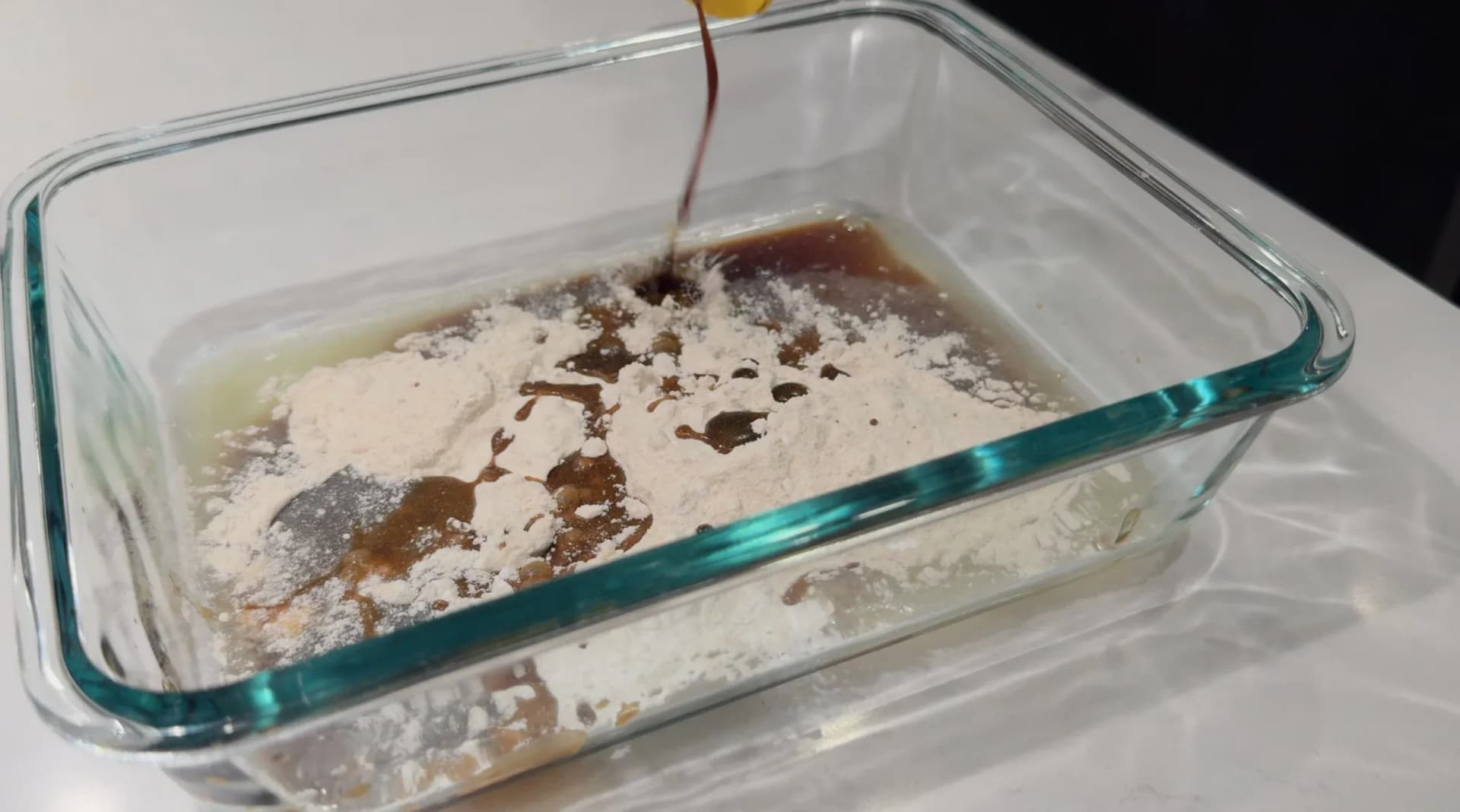 Dark liquid being poured into a glass baking dish containing white flour/starch mixture and other wet ingredients.