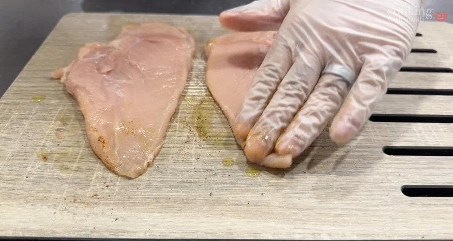 Two raw chicken breasts on a wooden cutting board being seasoned with salt and pepper from above while wearing food-safe gloves.