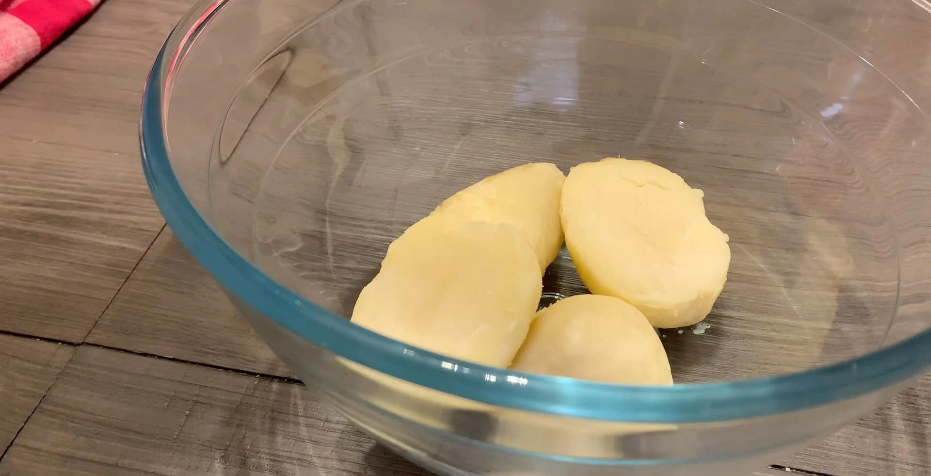 Three peeled potatoes in a clear glass bowl on a wooden surface.