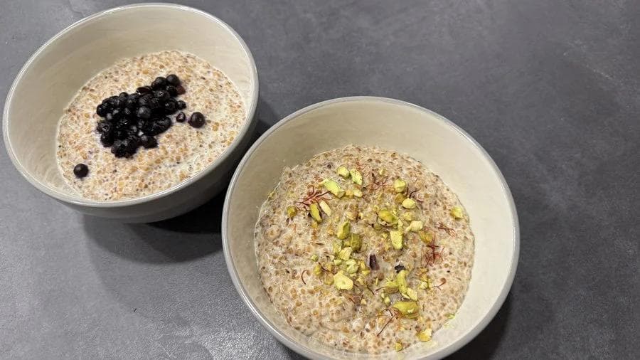 Two bowls of creamy cracked wheat porridge topped with blueberries and pistachios on gray surface.