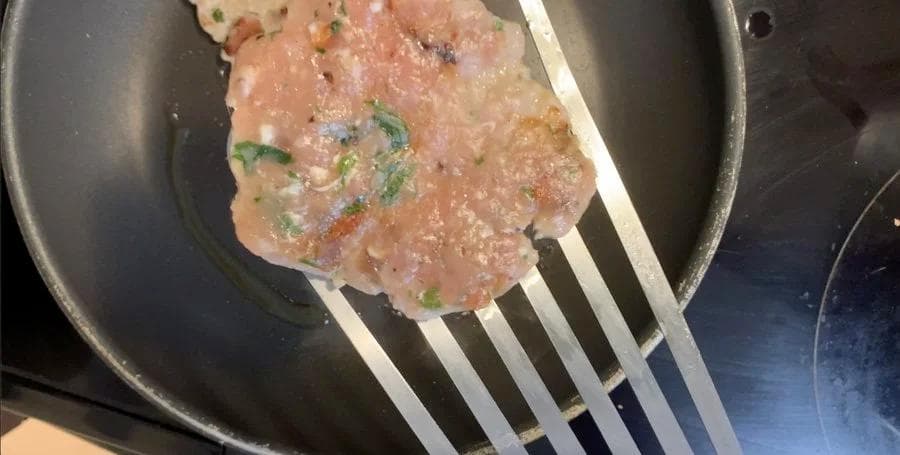 Sausage patty cooking in a skillet, being flipped with a spatula, topped with green onions and seasonings.