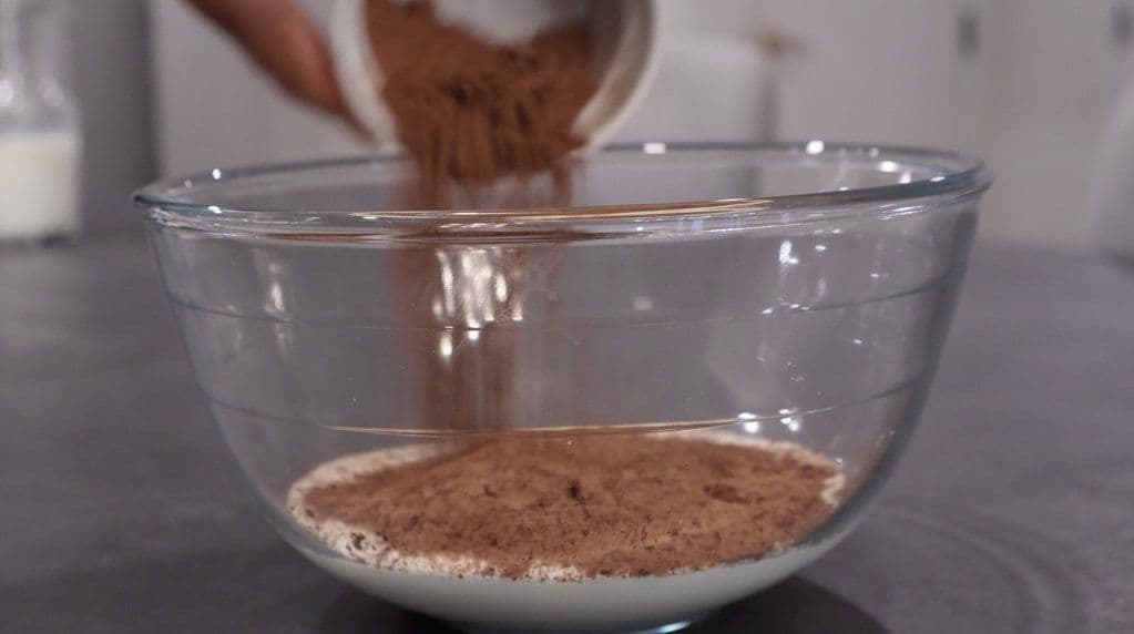 Cocoa powder being sifted into a glass bowl containing whipped cream mixture.