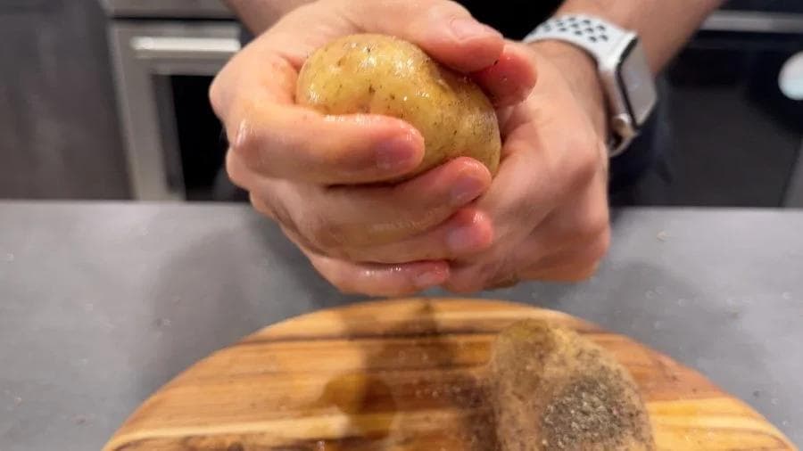 Hands rubbing a raw potato with olive oil and seasoning over a wooden board during preparation.