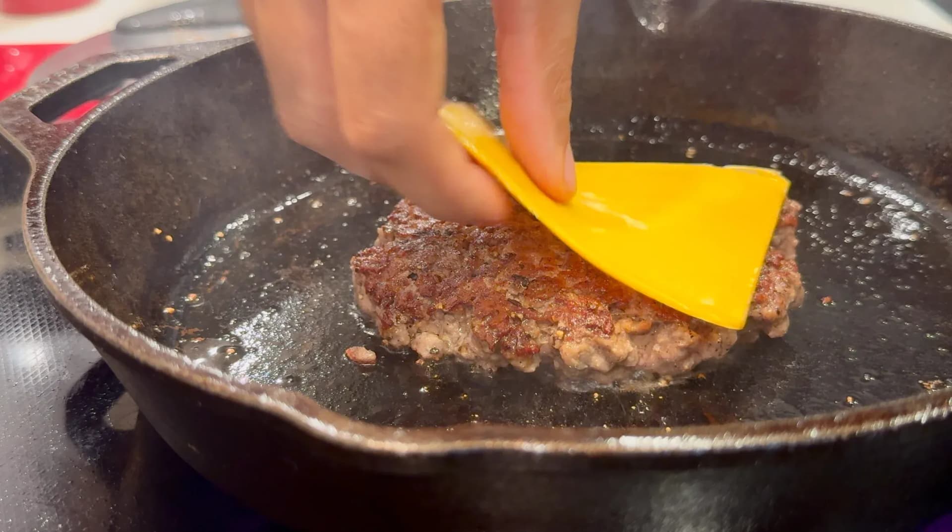 Flipping a cooked ground beef patty in a cast iron skillet with a yellow spatula.