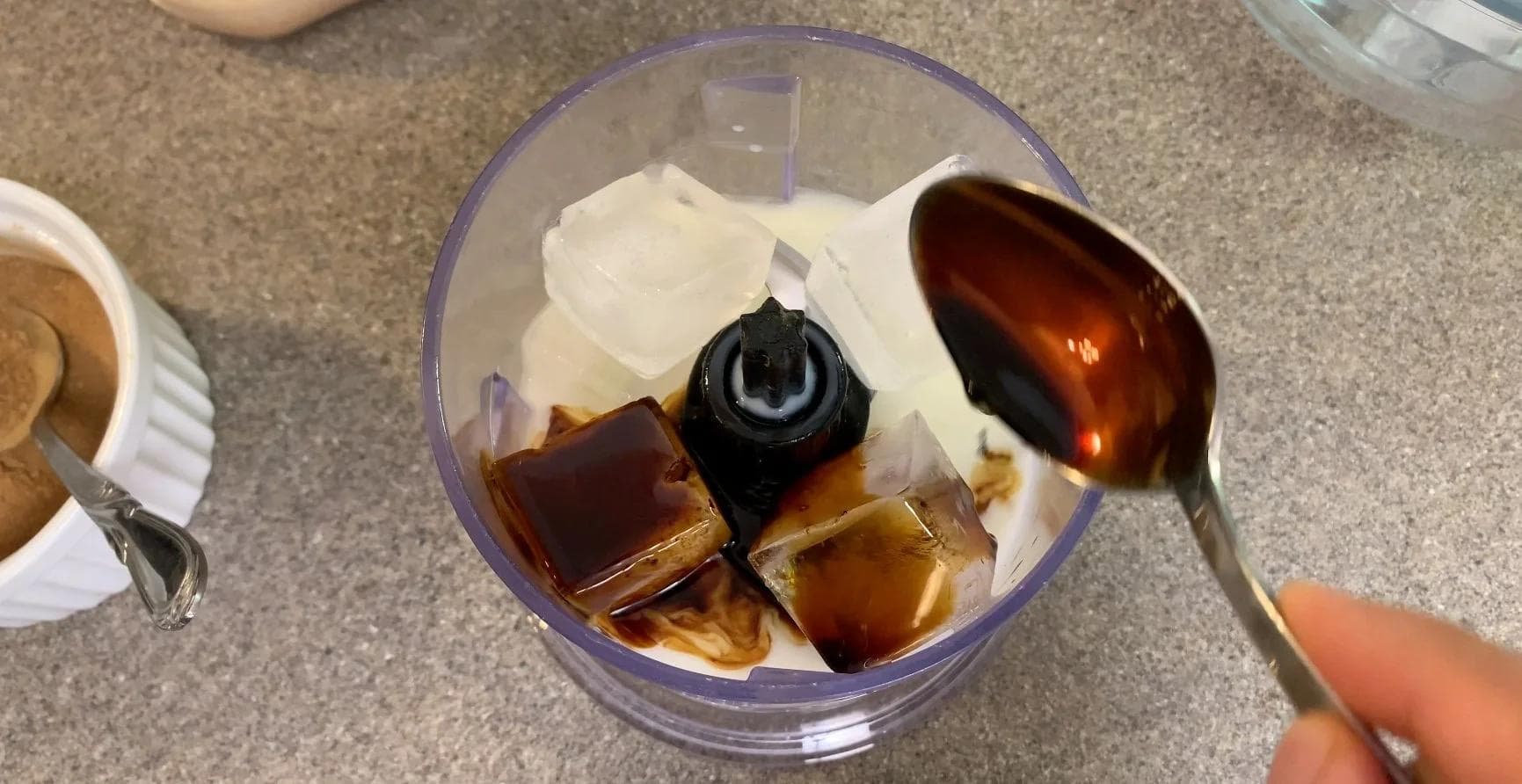 Pouring caramel-colored coffee mixture from a spoon into a blender containing ice and ingredients for pumpkin spice frappuccino.