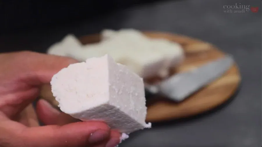 Hand holding a thick cube of paneer cheese with marinated paneer pieces on a wooden board in the background.