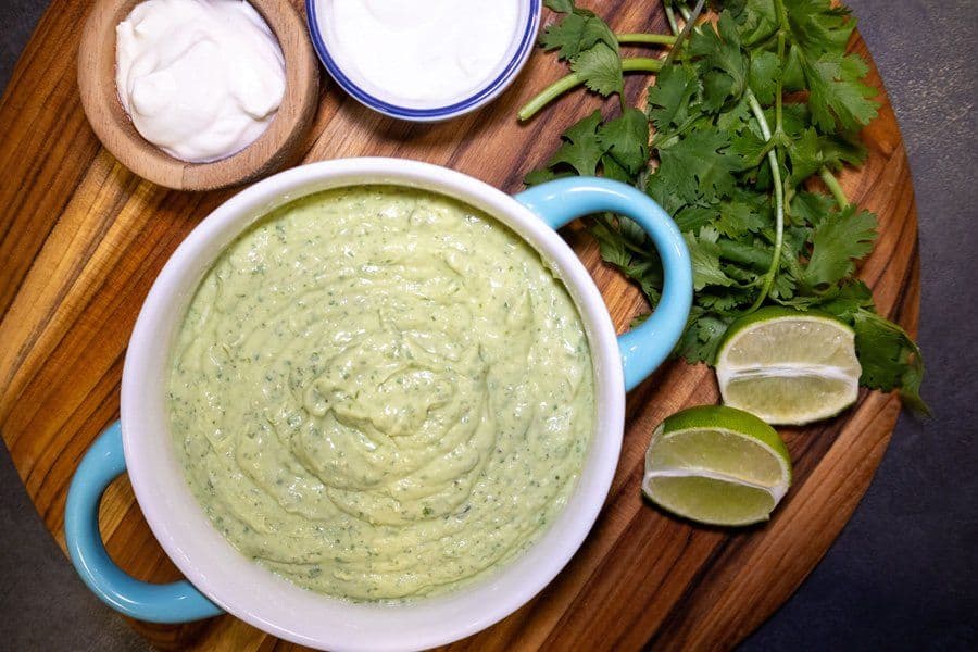 Creamy avocado lime crema in a white bowl with blue handles, surrounded by fresh cilantro and lime wedges on a wooden board.