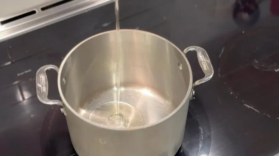 Oil being drizzled into an empty stainless steel pot on a stovetop.