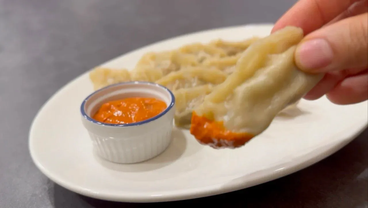 Hand dipping steamed chicken momo into orange chutney in a white ramekin, served on a plate.