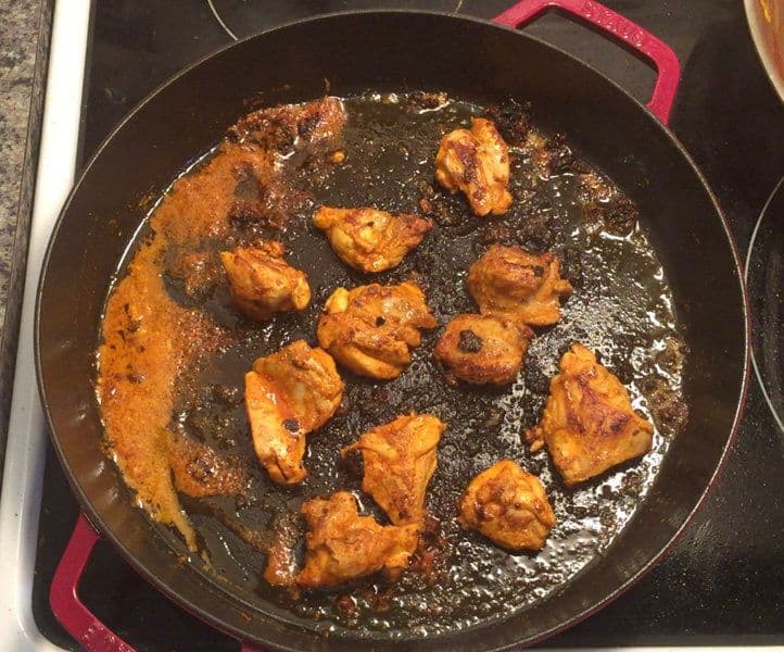 Chicken tikka pieces searing in a cast iron pan with caramelized marinade and oil.