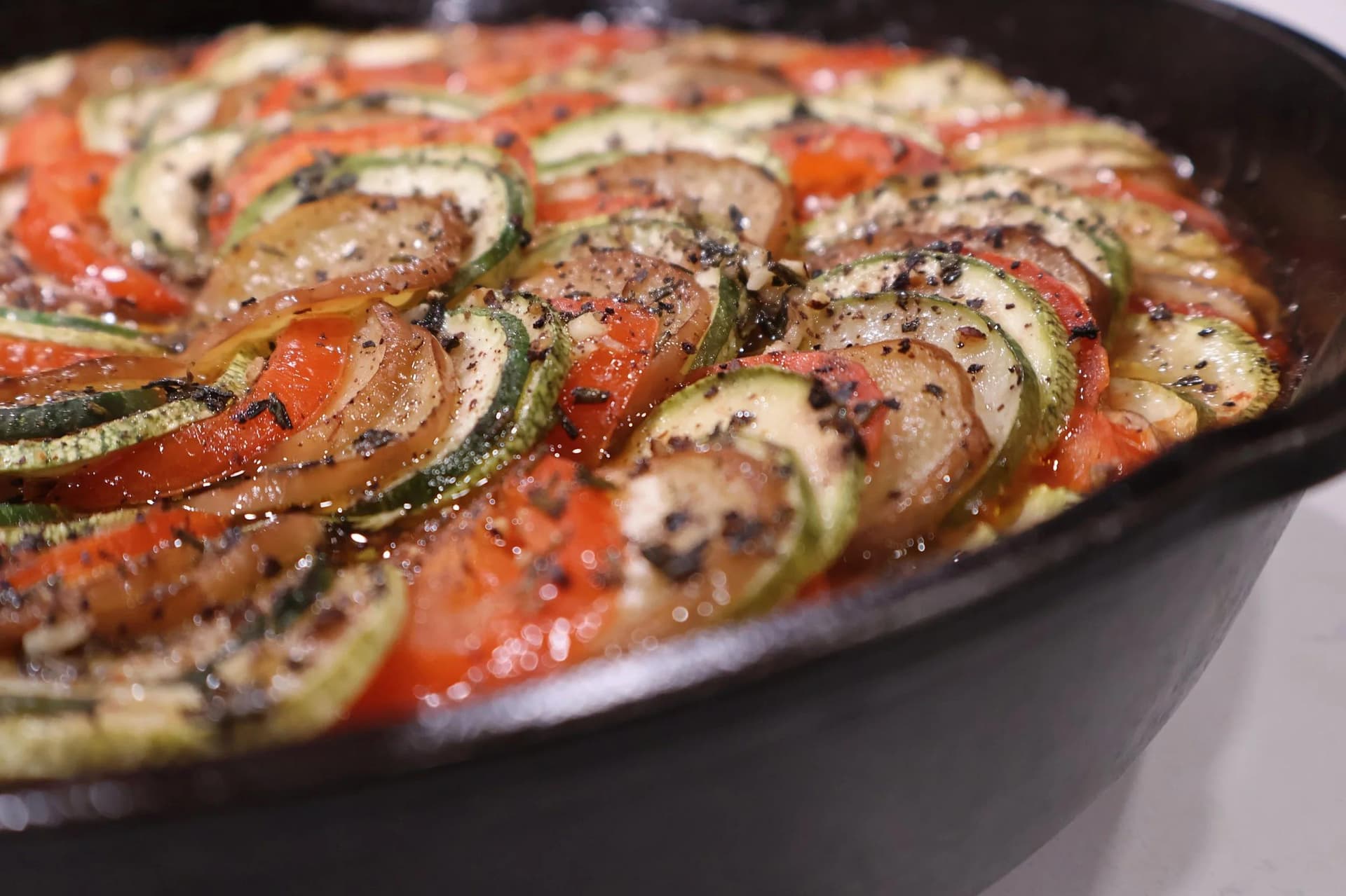 Sliced zucchini, tomatoes, and onions arranged in overlapping spiral pattern in a black baking dish, seasoned with herbs and pepper.