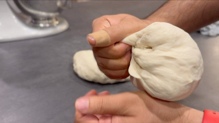 Hands folding pizza dough onto itself to create surface tension before the first rise.