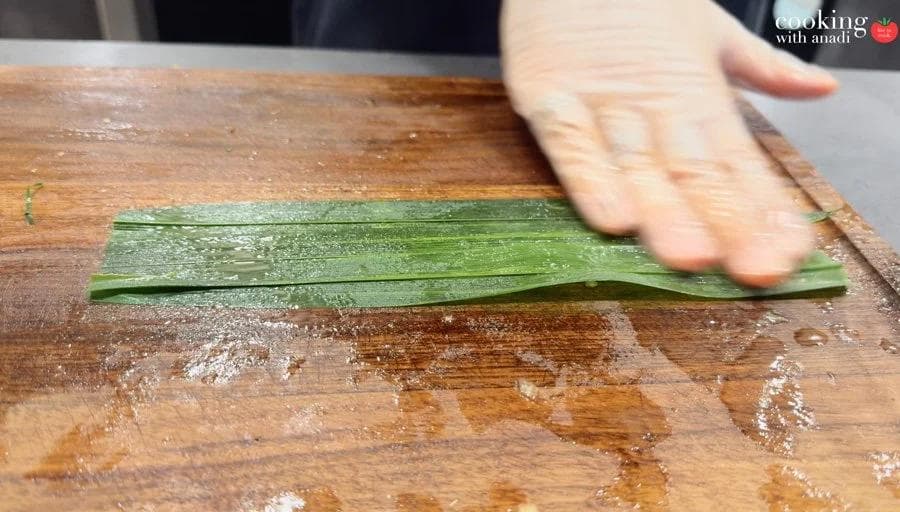 Hand pressing flat 2-3 pandan leaves on a wooden cutting board, leaves lightly oiled and neatly stacked