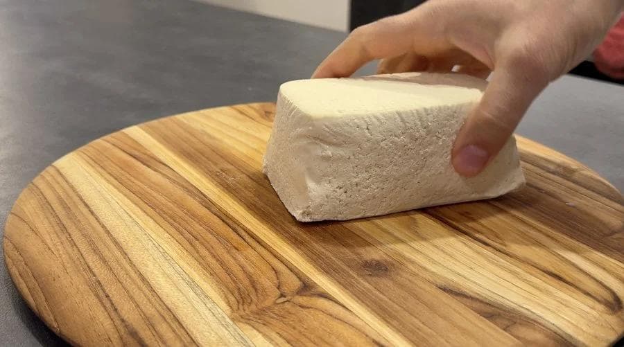 Hand patting tofu block dry with paper towel on wooden cutting board.