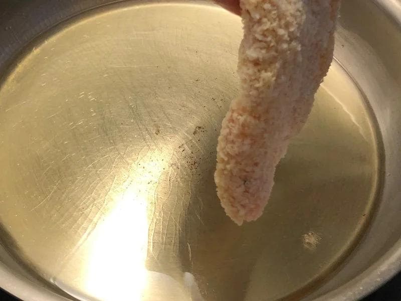 Panko-crusted chicken cutlet being fried in oil in a stainless steel pan until golden brown.