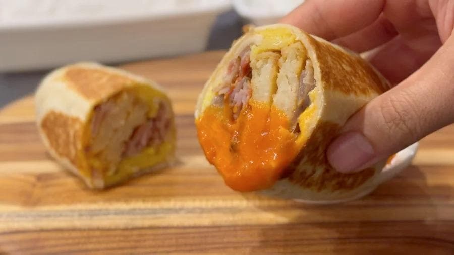 Hand holding a breakfast burrito cut in half, showing melted cheese and ham filling inside a golden tortilla.