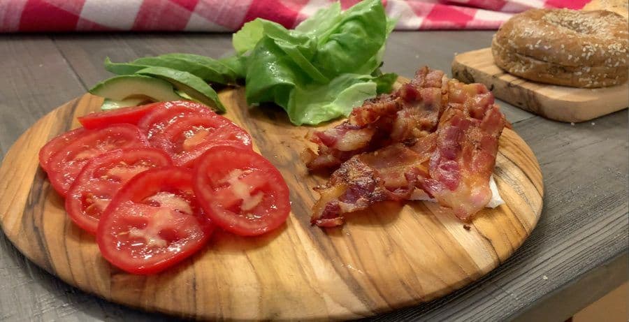 Wooden cutting board with sliced red tomatoes, fresh basil, cooked bacon strips, and whole wheat bagel.