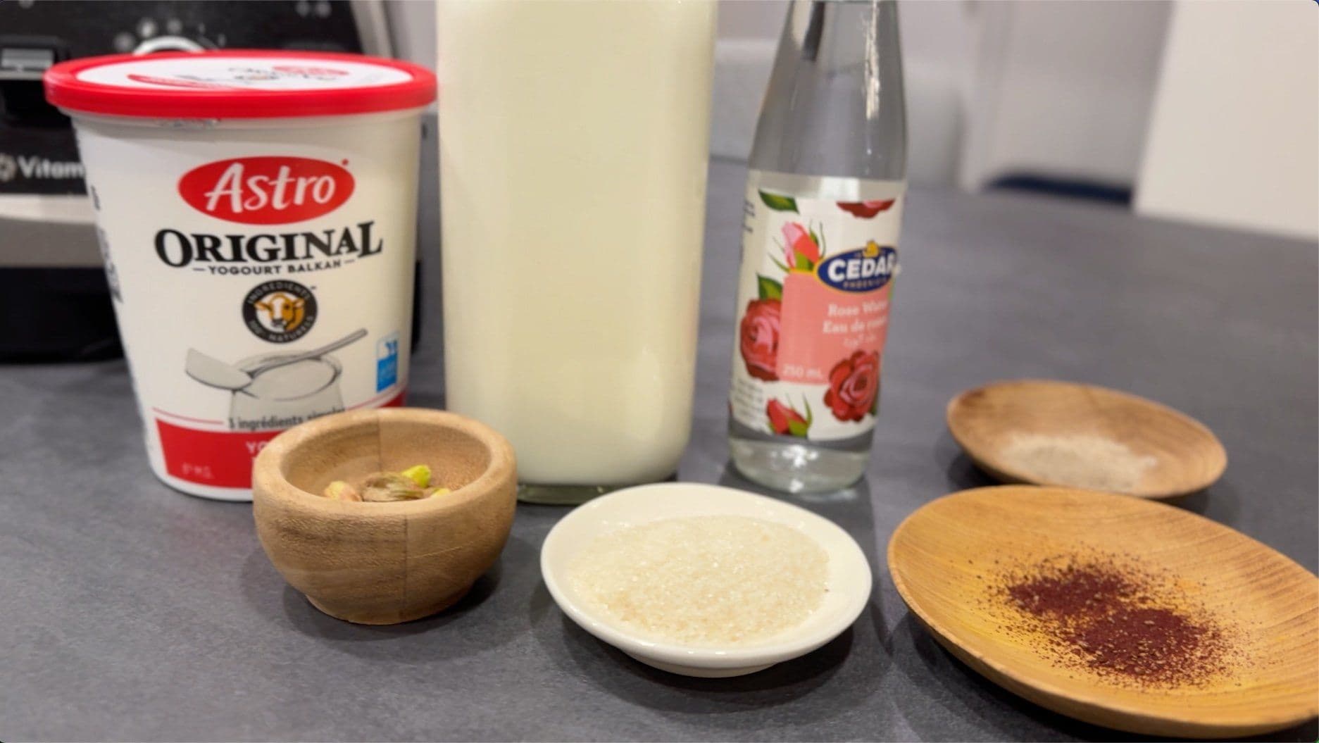Ingredients for Kesar Pista Lassi displayed on gray surface: yogurt, milk, saffron threads, pistachios, and rose water.