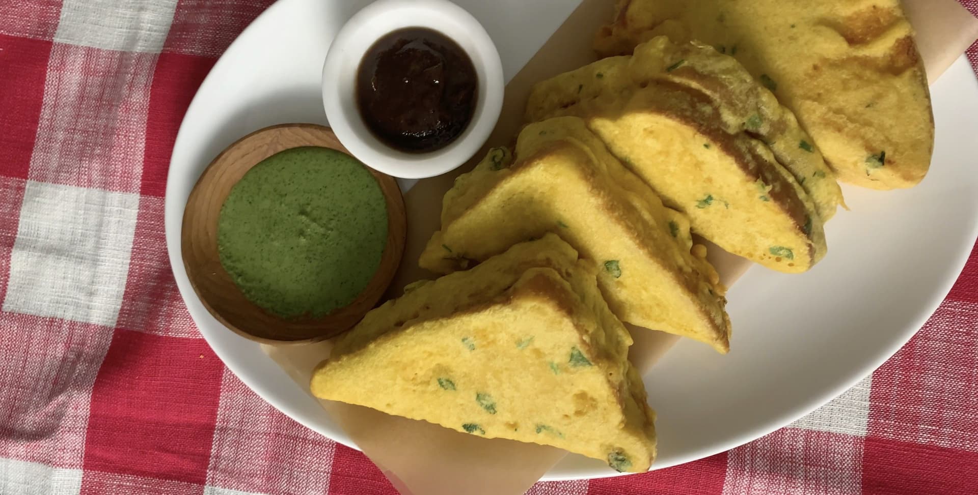 Plate of golden aloo bread pakora with green chutney and tamarind sauce on a checkered tablecloth.