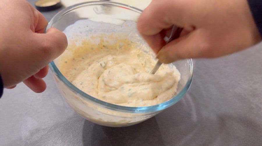 Hands stirring creamy beige ranch dip with visible herbs in a glass bowl.