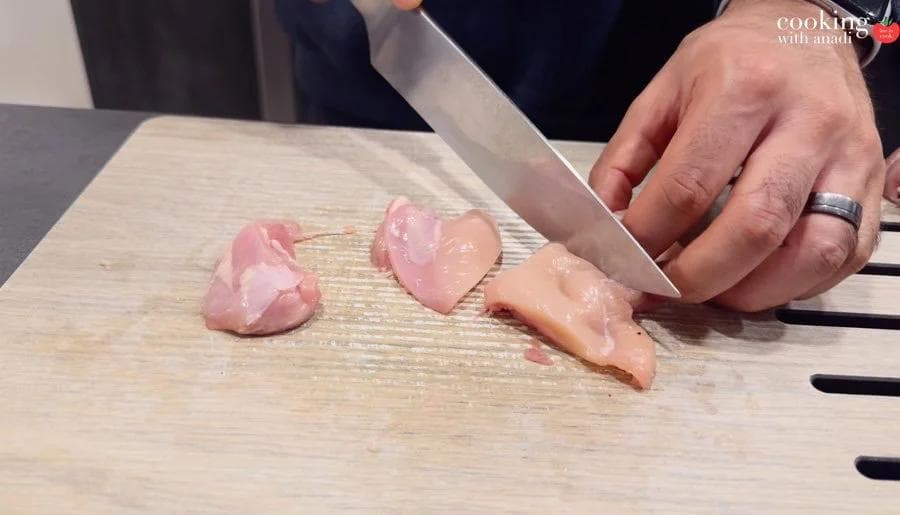 Hands cutting raw chicken thighs into bite-sized pieces on a wooden cutting board with a chef's knife.