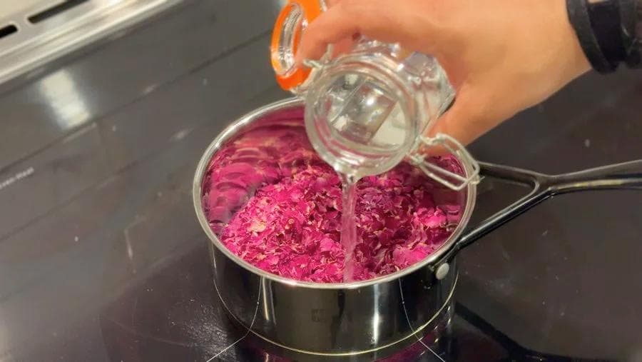 Pouring rose petal infused water from a glass measuring cup into a saucepan of deep pink rose petals.