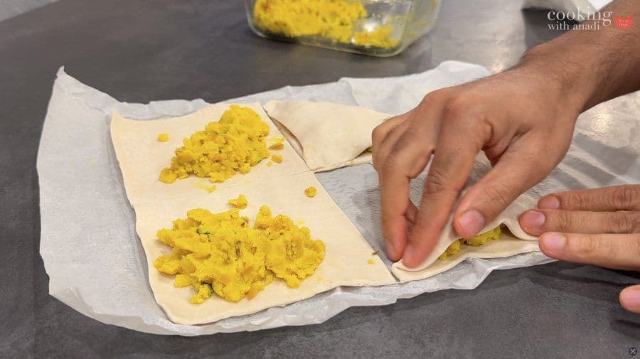 Folding puff pastry squares diagonally over yellow potato filling to form turnovers on parchment paper.