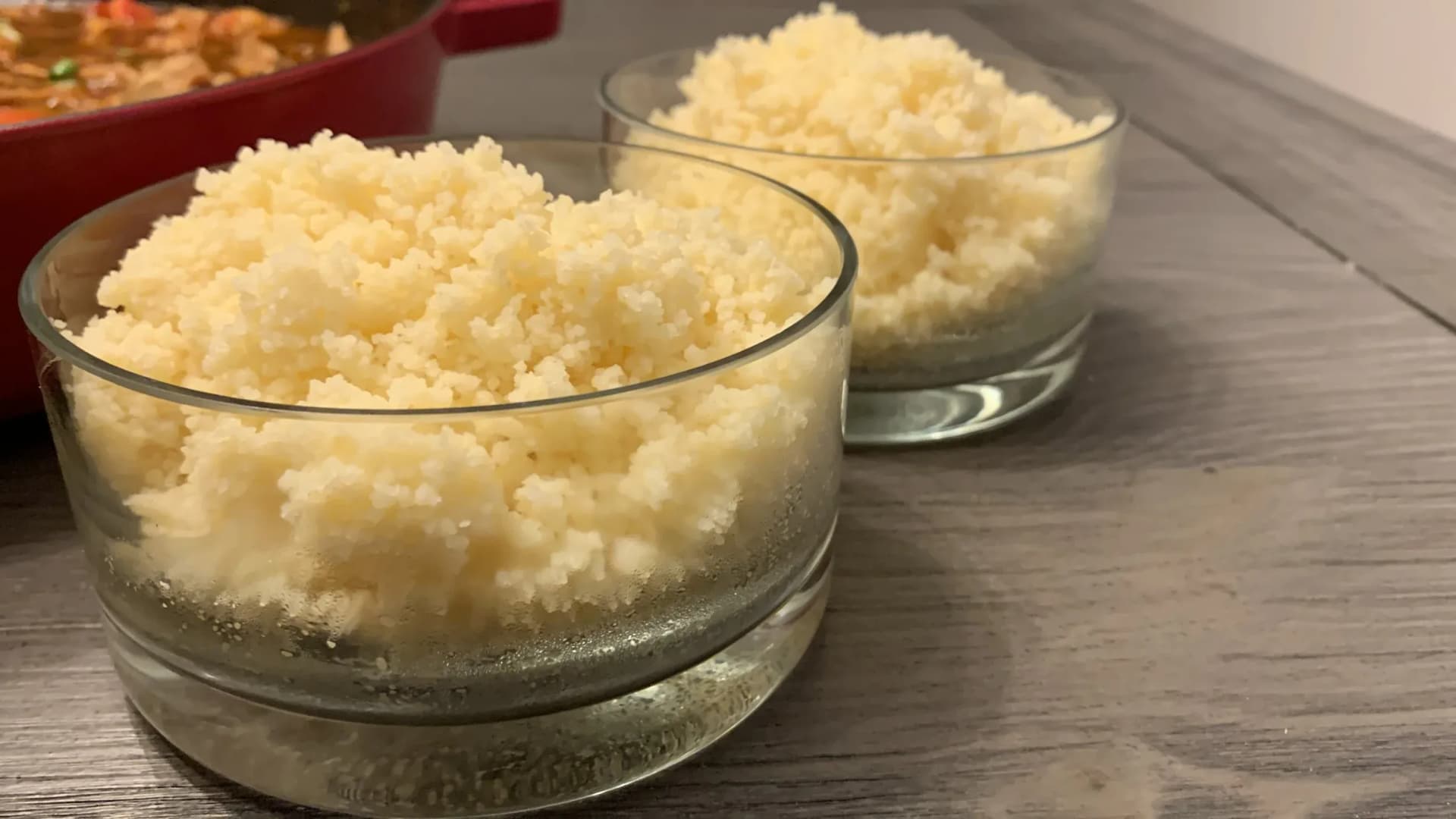 Two glasses filled with fluffy couscous, displayed on a wooden surface with a red cooking pot in the background.