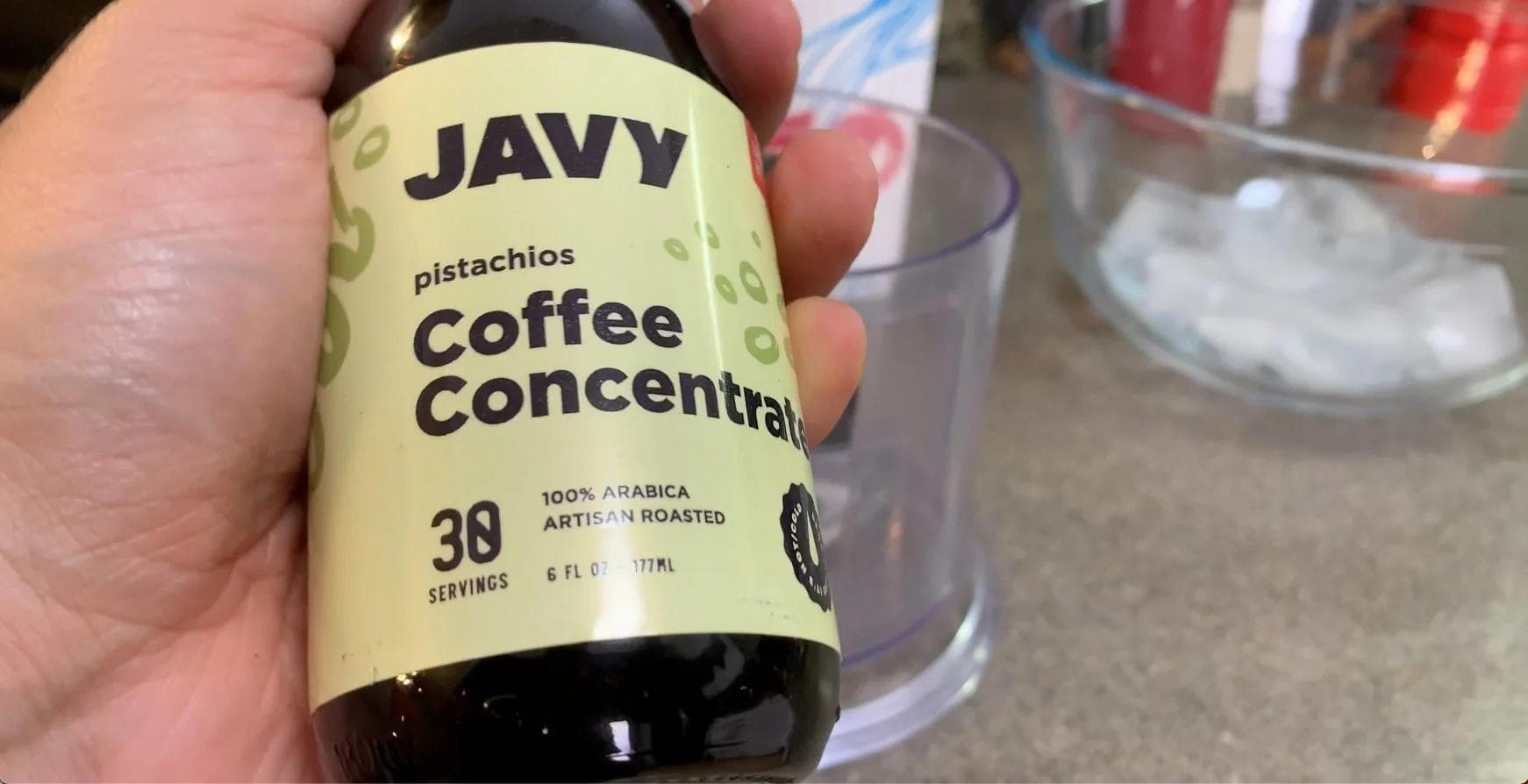 Hand holding a bottle of Javy pistachio coffee concentrate next to a glass with ice.
