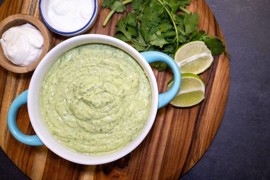 Creamy avocado lime crema in a white bowl with cilantro, lime wedges, and sour cream on a wooden board.