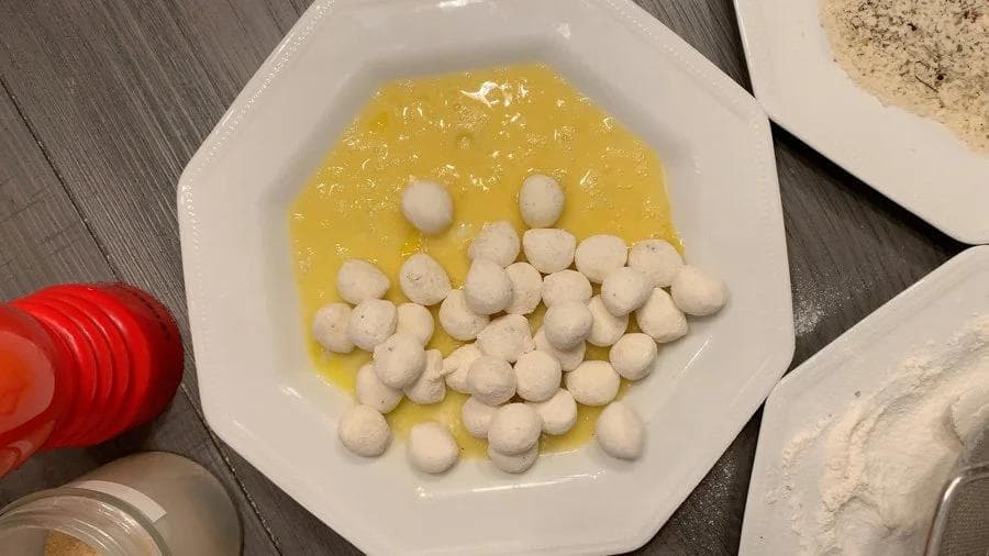 Coated mozzarella balls in egg wash on a white plate, ready for breadcrumb coating.