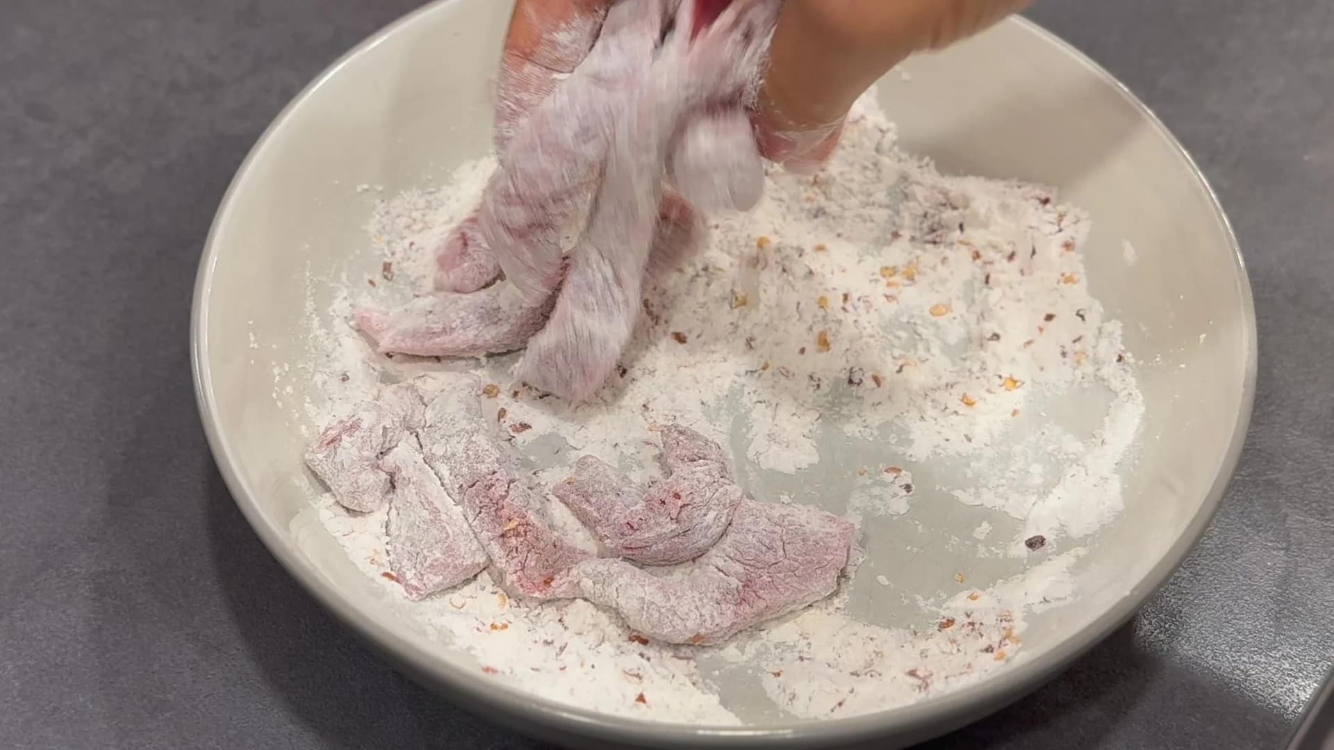 Raw beef stir fry strips being coated in seasoned flour mixture in a white bowl.