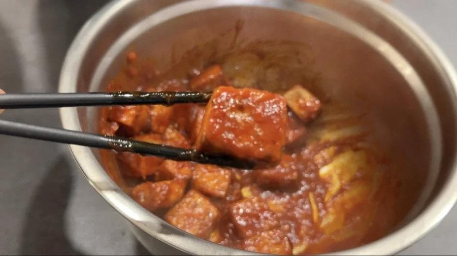 Chopsticks lifting a piece of glazed tofu from a bowl of glossy brown sauce with coated tofu pieces.