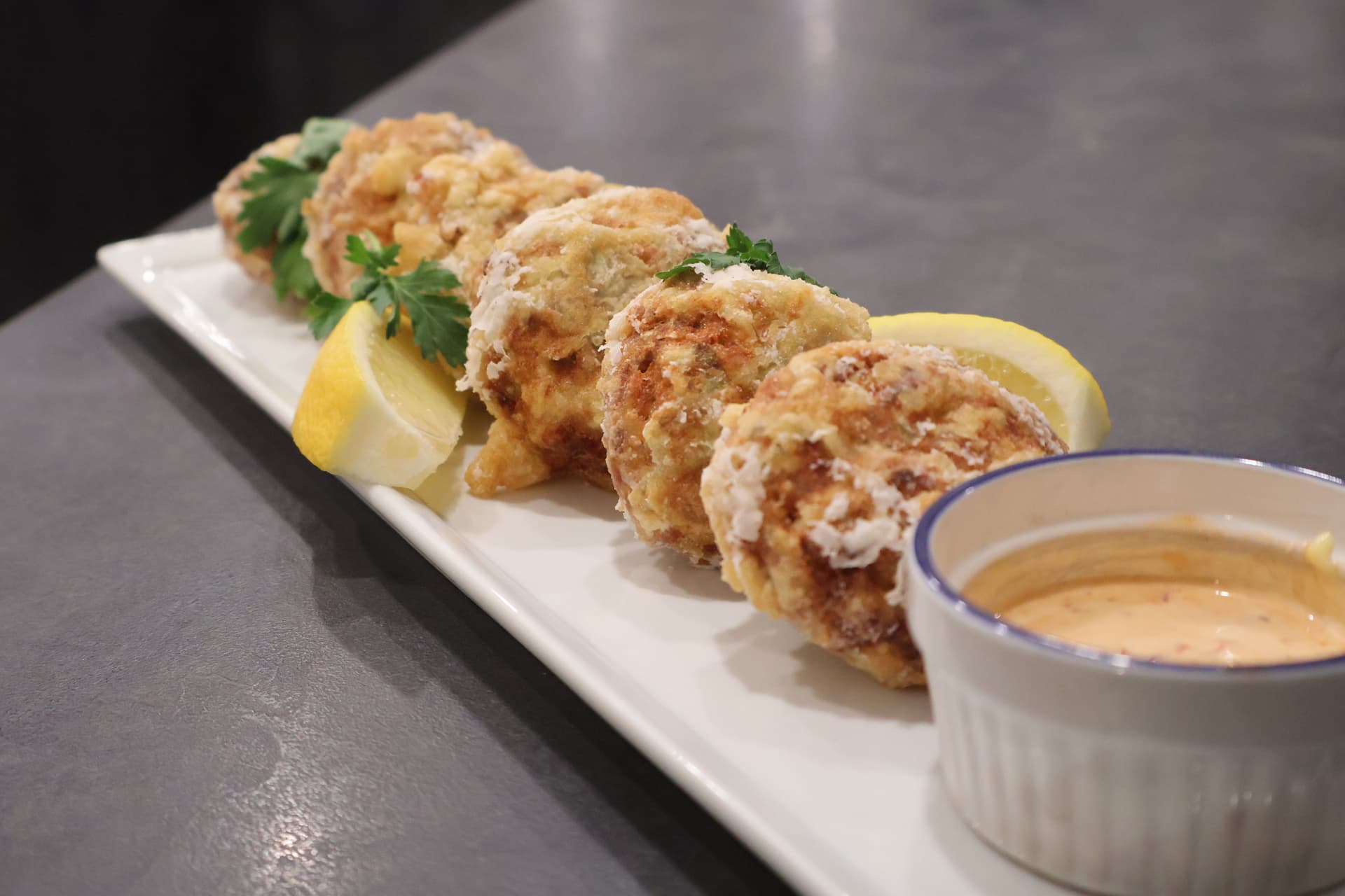 Golden-brown perkedel daging arranged on white plate with lemon wedges, parsley, and creamy dipping sauce.