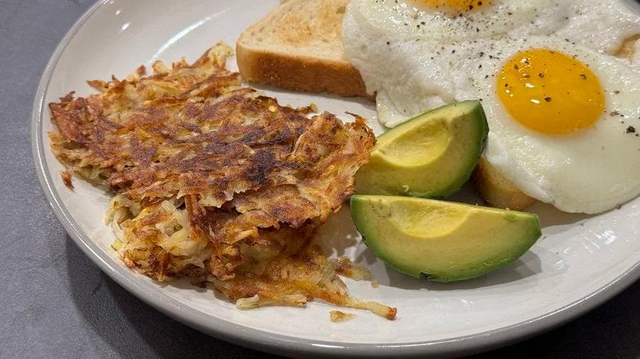 Crispy golden hash browns served on a white plate with fried eggs, avocado slices, and toasted bread.