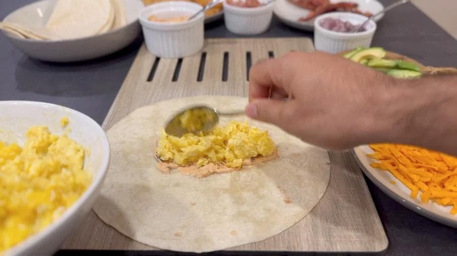 Assembling a breakfast burrito with scrambled eggs, ham, and pickles on a flour tortilla with additional fillings nearby.