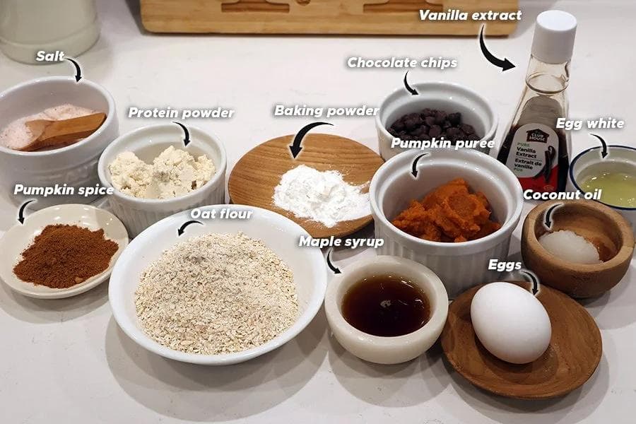Ingredients for high protein pumpkin chocolate chip cookies laid out in bowls and wooden dishes on a white surface.